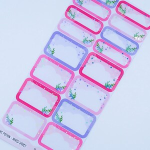 Puede incluir: Una hoja de pegatinas rectangulares con bordes rosas y morados. Cada pegatina tiene un fondo similar a una acuarela y está decorada con pequeños detalles florales y de estrellas. El texto "LOVE POTION - MIXED BOXES" y una firma son visibles.