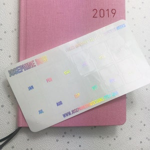 May include: A pink 2019 planner with a holographic sticker sheet of monthly tabs. The sticker sheet has the text "FOILED MONTHLY TABS FOR HOBONICHI WEEKS" and the months of the year. The website address is also visible.
