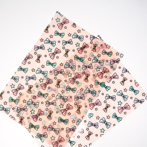 Può includere: Diversi fogli di carta fantasia con un motivo ripetuto di fiocchi e stelle colorate su sfondo pesca. I fiocchi sono bordati di nero e riempiti di colori pastello. Le stelle sono bianche.