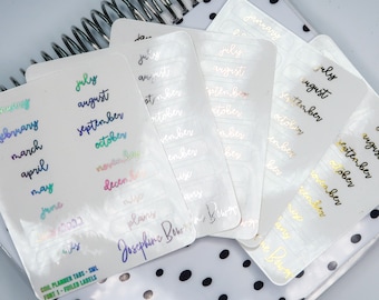 Puede incluir: Un conjunto de pestañas de planificador transparentes e iridiscentes con letras doradas. Las pestañas presentan los meses del año, incluyendo enero, febrero, marzo, abril, mayo, junio, julio, agosto, septiembre, octubre, noviembre y diciembre.