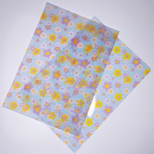 Könnte beinhalten: Zwei Blätter durchscheinendes Papier mit hellblauem Hintergrund und einem sich wiederholenden Muster aus gelben und violetten Blumen.