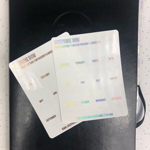 May include: Two sheets of clear, iridescent stickers with the text "Josephine Bow" and "Full Color Monthly Tabs for Passion Planner" printed on them. The stickers are labeled with the months of the year in rainbow colors.