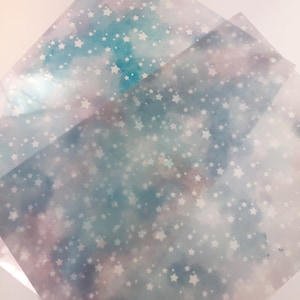 May include: Two sheets of translucent paper with a watercolor background in shades of blue, pink, and purple. The paper is covered in a repeating pattern of white stars.