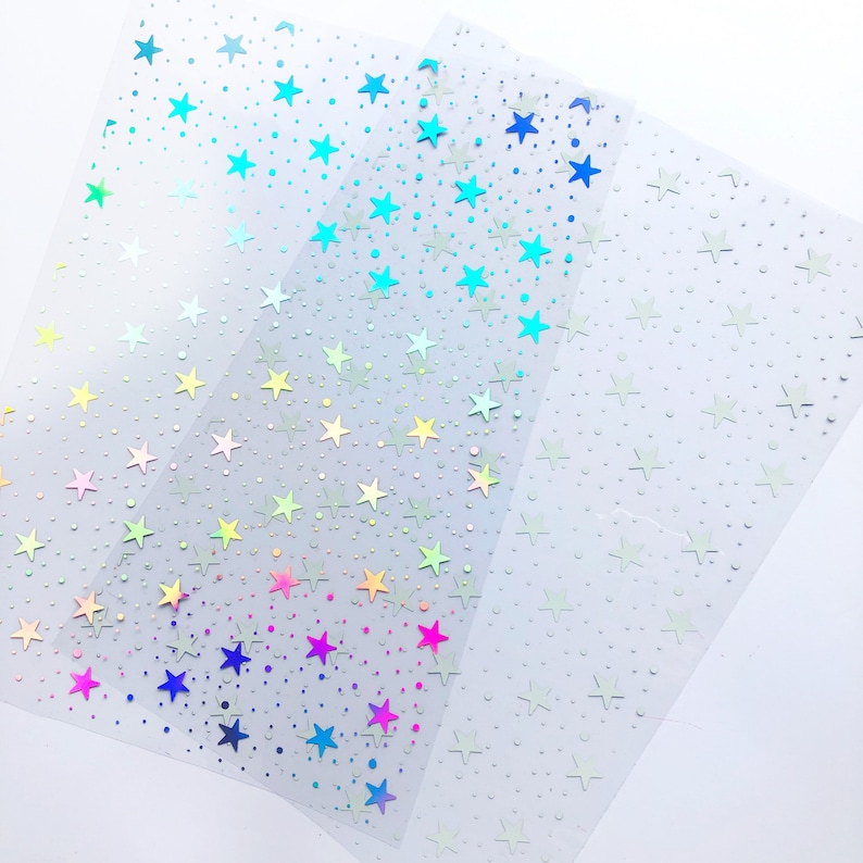 May include: Three sheets of clear, iridescent vinyl with a repeating pattern of stars and small dots. The stars are in a gradient of colors, starting with blue at the top and fading to pink at the bottom. The sheets are stacked on top of each other, with the blue sheet on top and the pink sheet on the bottom.