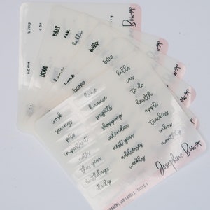 May include: A set of clear plastic tab labels with black handwritten text. The labels are for a binder or planner and include words like "home", "bills", "work", "savings", "finance", "projects", "shopping", "calendar", "to do", "health", "appts", "trackers", "inbox", "monthly", "next year", "this year", "important", "notes", "birthdays", "addresses", "weekly", "daily", and "pets". The labels are from Josephine Bows.