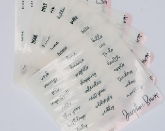 Puede incluir: Un conjunto de etiquetas de pestañas de plástico transparente con texto escrito a mano en negro. Las etiquetas son para una carpeta o un planificador e incluyen palabras como "hogar", "facturas", "trabajo", "ahorros", "finanzas", "proyectos", "compras", "calendario", "que hacer", "salud", "citas", "seguimiento", "bandeja de entrada", "mensual", "próximo año", "este año", "importante", "notas", "cumpleaños", "direcciones", "semanal", "diario", y "mascotas". Las etiquetas son de Josephine Bows.