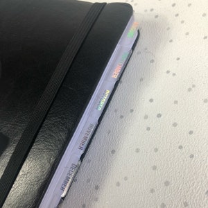 May include: A black leather-bound planner with a black elastic band closure. The planner has iridescent tabs for each month, starting with December and ending with August.