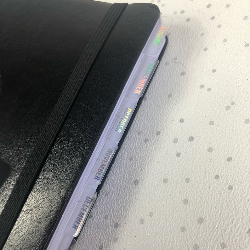May include: A black leather-bound planner with a black elastic band closure. The planner has iridescent tabs for each month, starting with December and ending with August.