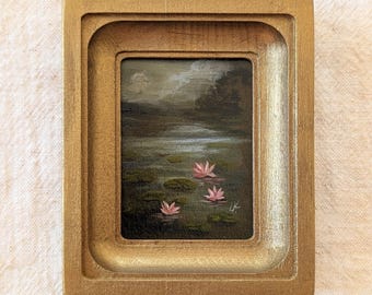 May include: A small rectangular painting in a thick gold-toned frame depicting a dark pond with three pink water lilies floating on the surface. The scene features a moody, overcast sky above a shadowed landscape with trees in the background.