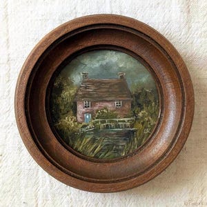 以下が含まれることがあります： 茶色の屋根と明るい壁のコテージが描かれた小さな油絵が入った円形の木製フレーム。緑の茂みと暗く曇った空が広がる水辺の風景。右下にLKのイニシャルが描かれています。