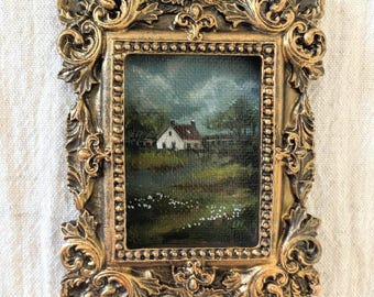 May include: A small rectangular painting of a white house with a red roof in a green field under a cloudy sky, encased in an ornate, metallic gold-colored frame with intricate scrollwork and a beaded inner border detail.