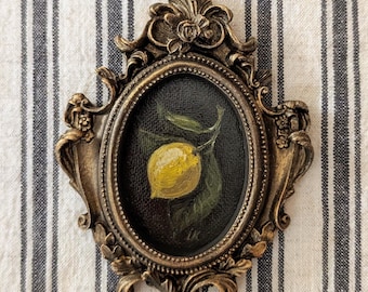 May include: An oval-shaped painting of a yellow lemon with a green leaf on a dark background, encased in an ornate, metallic bronze-coloured frame with intricate floral and scrollwork detailing. The artist's initials, LK, are visible at the bottom.