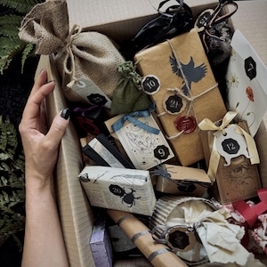 Peut inclure: Une boîte en carton remplie de petits cadeaux emballés dans du papier kraft, de la toile de jute et du papier à motifs. On y trouve un sac en jute, un paquet avec une silhouette d'oiseau noir et un sceau en cire rouge, et des étiquettes numérotées.
