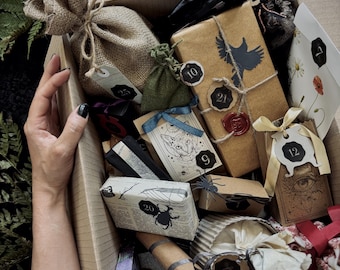Puede incluir: Una caja de cartón llena de pequeños regalos envueltos en papel marrón, arpillera y papel estampado. Incluye un saco de arpillera, un paquete con la silueta de un pájaro negro y un sello de cera roja, una caja con ilustración de gato y etiquetas numeradas.