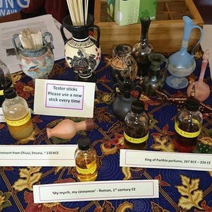May include: A display of ancient perfume bottles and tester sticks on a patterned tablecloth. The bottles vary in size and shape, some with yellow labels. Signs indicate the perfume's origin and era, including "My myrrh, my cinnamon" from Roman times.