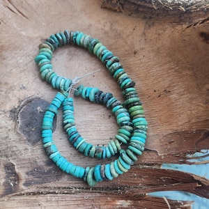May include: A strand of turquoise beads, with a mix of blue and green tones, arranged in a circular shape on a wooden surface.
