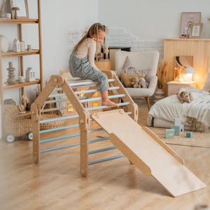 May include: A wooden climbing frame with a slide for children. The frame is made of natural wood and has a light blue and white color scheme. A child is climbing up the frame.