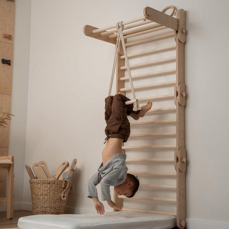May include: A child hangs upside down from a wooden swing set with a rope. The swing set is attached to a wall and has a ladder. The child is wearing a gray shirt and brown pants.