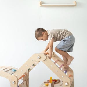 May include: A child climbs a wooden play structure with colorful rungs. The structure is made of natural wood and has a curved ramp and a ladder.