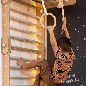 May include: A child hangs from wooden gym rings attached to a wooden climbing wall with a string of lights. The wall has a light wood finish and the child is wearing a brown t-shirt with a black and white whale print. The wall is against a dark blue background with white dots.