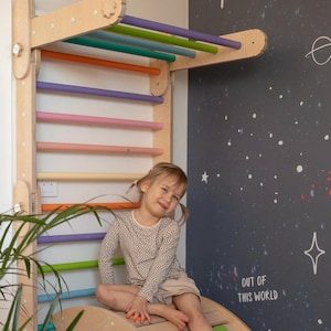 May include: A young child sits on a wooden rocker, facing the camera and smiling. The child is wearing a beige long-sleeved shirt with black polka dots and beige shorts. Behind the child is a wooden climbing wall with colorful wooden rungs. The wall is mounted on a white wall with a black chalkboard painted on the right side. The chalkboard has white stars and the text "Out of this world" written on it.