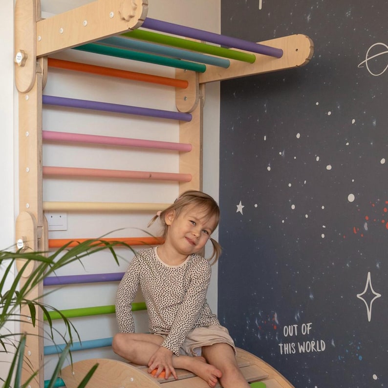 May include: A young child sits on a wooden rocker, facing the camera and smiling. The child is wearing a beige long-sleeved shirt with black polka dots and beige shorts. Behind the child is a wooden climbing wall with colorful wooden rungs. The wall is mounted on a white wall with a black chalkboard painted on the right side. The chalkboard has white stars and the text "Out of this world" written on it.