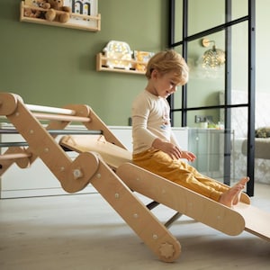 May include: A child sits on a wooden slide attached to a climbing frame. The slide is made of natural wood and has a rounded edge. The climbing frame has a ladder and a rope.