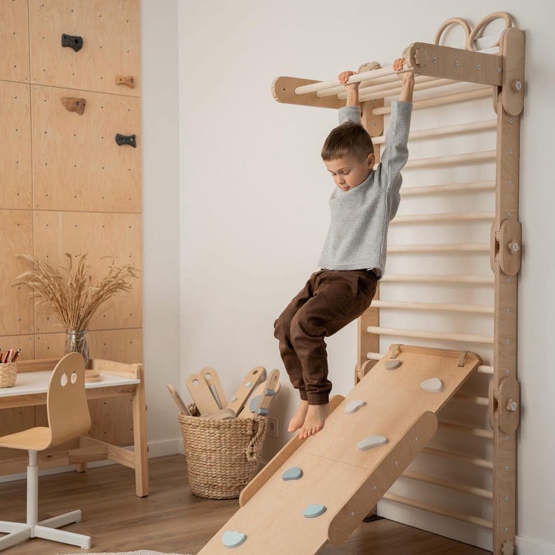 May include: A wooden indoor play structure with a climbing wall, monkey bars, and a slide. A child is hanging from the monkey bars.