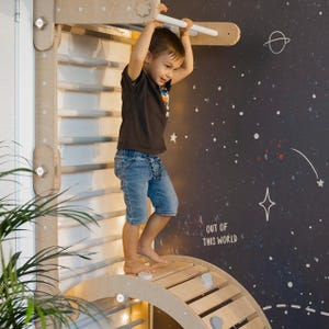 May include: A child climbs on a wooden indoor play structure with a climbing wall, a curved ramp, and a horizontal bar. The wall behind the structure is painted dark blue with white stars and the text "Out of this world".