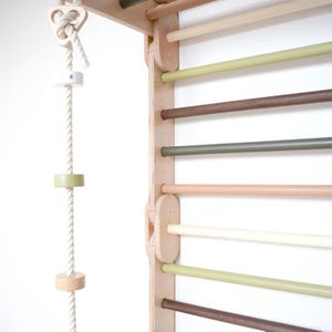 May include: A wooden climbing wall with a variety of coloured bars. The wall is made of light wood and the bars are in shades of brown, green, and beige. A rope with wooden rings hangs from the top of the wall.