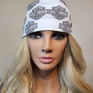 May include: A mannequin wearing a white Harley-Davidson bandana with black text and logos. The mannequin also has long blonde hair, a silver necklace, and a large Harley-Davidson pendant.