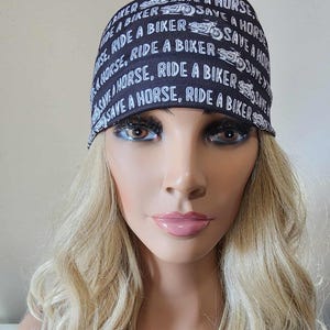 May include: A black bandana with white text that reads "SAVE A HORSE, RIDE A BIKER" is worn on a mannequin head with long blonde hair. A silver necklace with a Harley-Davidson logo pendant is also visible.