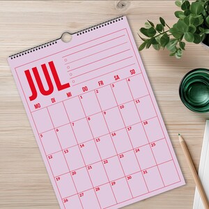 Pu&ograve; includere: Un calendario da parete rosa per luglio con testo rosso e linee a griglia. Il calendario mostra i giorni della settimana e le date numerate. La parola "JUL" &egrave; ben visibile in alto.