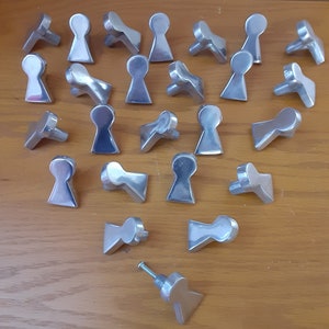May include: A collection of silver-colored keyhole-shaped cabinet knobs. The knobs are made of metal and have a smooth, reflective surface. One knob has a screw attached. The knobs are arranged on a wooden surface.