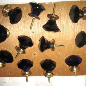 May include: A collection of eleven cabinet knobs featuring faceted, deep blue glass heads attached to circular brass-toned metal bases. Each knob includes a threaded metal screw extending from the back for installation on furniture or cabinetry.