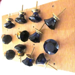 May include: A collection of eleven faceted dark blue glass cabinet knobs with brass-toned metal bases and threaded mounting screws. Each knob features a multi-faceted geometric shape that reflects light, arranged on a light brown wooden board.
