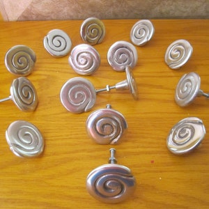 May include: A collection of silver-toned cabinet knobs with a spiral design. The knobs are arranged on a wooden surface, showcasing their reflective quality and unique shape. Each knob has a screw for easy installation.