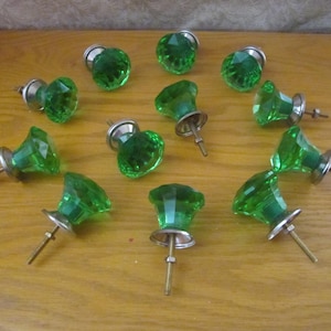 May include: A collection of twelve green glass cabinet knobs with faceted surfaces and silver-toned metal bases. The knobs are arranged on a light brown wooden surface, showcasing their decorative design and potential for furniture upgrades.