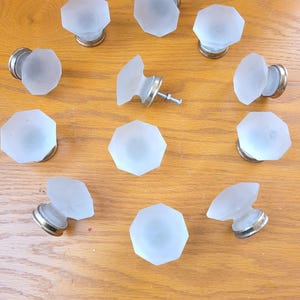 May include: A collection of frosted glass cabinet knobs with an octagonal design. Each knob has a silver-colored base and a screw for installation. The knobs are arranged on a wooden surface.