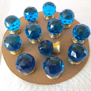 May include: A collection of twelve faceted, blue glass cabinet knobs. Each knob has a round, jewel-like appearance and is mounted on a gold-colored base. The knobs are arranged on a brown, circular surface.