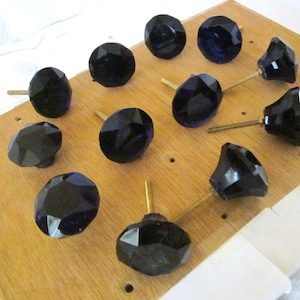 May include: A collection of faceted, dark blue glass cabinet knobs with brass stems. The knobs are arranged on a light brown wooden surface, showcasing their geometric shapes and reflective qualities. Ideal for furniture restoration or DIY projects.