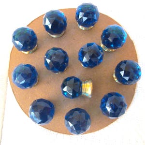 May include: A collection of twelve faceted, deep blue glass knobs with brass-colored bases. The knobs are arranged in a circular pattern on a brown cardboard surface, showcasing their jewel-like appearance.