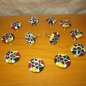 May include: A collection of eleven round cabinet knobs featuring a multi-colored floral pattern in red, yellow, green, and white. Each knob has a smooth, glossy finish and is attached to a metallic base, arranged in a circular formation.