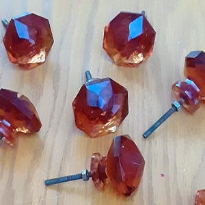 May include: A collection of faceted, amber-colored glass cabinet knobs. Each knob has a unique, geometric design and is attached to a metal screw for easy installation. The knobs are arranged on a light-colored wooden surface.