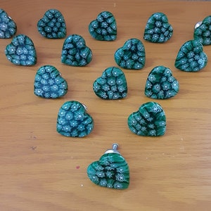 May include: A collection of heart-shaped cabinet knobs. Each knob is a vibrant green with a floral pattern in white and silver. The knobs are arranged on a light brown wooden surface, showcasing their decorative design.