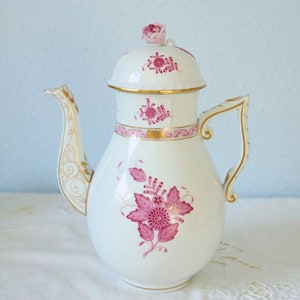 Op de afbeelding: Een witte porseleinen koffiepot met een roze bloemmotief, gouden randen en een sculpturale rozenknop op het deksel. Het lichaam toont een centraal magenta bloemenboeket, terwijl het handvat en de tuit zijn voorzien van gouden details.