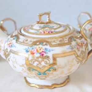 Op de afbeelding: Een wit porseleinen theeservies met gouden randen en bloemmotieven. De set bestaat uit een theepot, een suikerpot en een melkkan. De theepot heeft een deksel en een gebogen tuit. Het bloemmotief bevat roze, blauwe en gele bloemen.