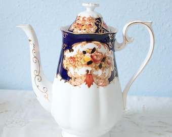 Pode incluir: Um bule de porcelana com base branca e seção superior azul profundo, decorado com filigrana dourada e um buquê floral central de rosas laranja, amarelas e cor-de-rosa. O bule inclui uma tampa correspondente com detalhes dourados.