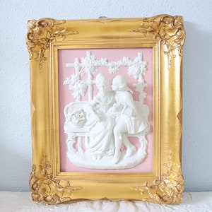 Puede incluir: Escultura en relieve enmarcada que representa una escena romántica con dos figuras, una mujer y un hombre, sentados en un banco. El relieve es blanco sobre un fondo rosa, enmarcado por un marco dorado ornamentado. El marco mide aproximadamente 30 cm por 25 cm.