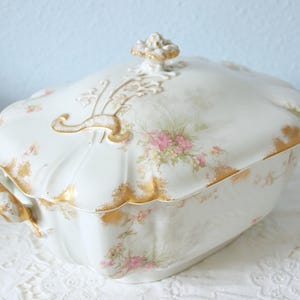 以下が含まれることがあります： ピンクと緑の花柄が施された、蓋付きの白い磁器製サービングディッシュ。縁と装飾は金でトリミングされています。ディッシュはスカラップの形をしており、蓋には装飾的なハンドルが付いています。